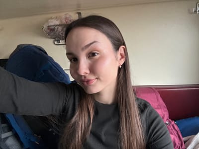 The image captures a selfie of a female individual inside what is consistent with a Russian train compartment, with the contextual location identified as Usolye-Sibirskoye, Russia. The subject is a young adult female, positioned centrally, looking directly into the camera with a neutral expression and a slight upward curve to her lips. She has long, straight, dark brown hair with lighter ends, parted down the middle, extending over her shoulders. She wears a dark, long-sleeved top. A small, clear-stone stud earring is visible in her right earlobe, along with a small ring in her right helix piercing.
To the subject's left, a large, dark blue backpack is visible. Above and behind her left shoulder, a metal wire rack is affixed to a light-colored wall, containing multiple translucent plastic bags. A silver metal hook is mounted higher on the wall to the right. To the subject's right, a padded maroon surface, consistent with a train seat or headrest, forms the background. Against this, a fuchsia-colored soft bag with a visible silver zipper is partially obscured by the subject. Below the fuchsia bag, a blue object and a white fabric item are partially visible. The immediate environment suggests a confined travel space within a passenger train car.