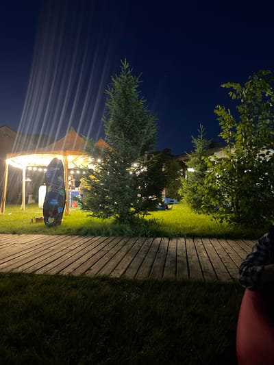 The image shows an outdoor scene at night, possibly in Pervomayskiye Prudy, Kazakhstan. There's a wooden deck or walkway on the right side, bordered by grass. A tent-like structure is illuminated with string lights, with a tree partially obscuring the view. An object, possibly a skateboard or sign with floral patterns and the word "2GIS" on it, is positioned in front of the tent. Above, the night sky is visible with the presence of trees. A person is seated on the upper right, with a portion of their checkered shirt visible.