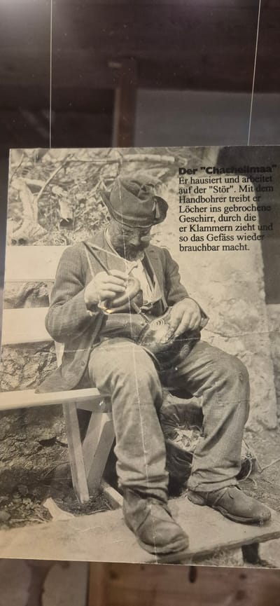 This black and white image features a close-up of a person dressed in what appears to be traditional or work attire. The person is seated on a simple wooden structure, possibly a bench or stool, and is engaged in a repair task. They are holding a hand drill and working on what looks like a piece of pottery or a ceramic vessel with several holes. The text next to the image, in German, indicates the person is known as "Der Chachelimaa" (The Pot Mender) and describes their work: they "lives and works on the 'Stör'. With the hand drill, he drills holes into the broken crockery, through which he pulls clamps and thus makes the vessel usable again." The setting appears to be outdoors, with a rocky or earthy background and some dry vegetation. The lighting suggests it could be daytime, and there are no obvious cues for weather conditions. The overall impression is of a craftsman diligently performing a repair.