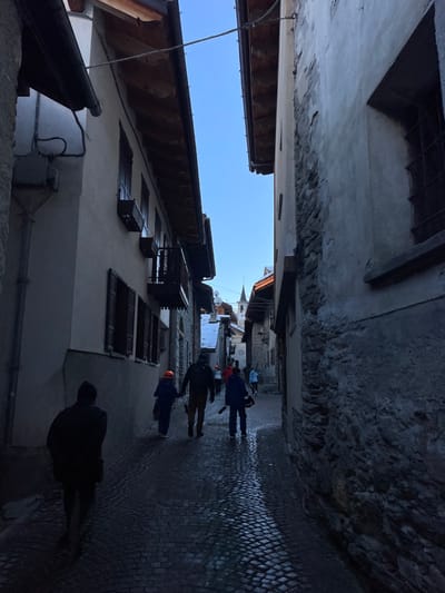 The image captures a narrow, shaded cobblestone street in Courmayeur, Italy, bordered by tall, historic buildings. The perspective is looking down the street, with pedestrians walking away from the viewer towards a distant church spire.

The street surface is paved with dark, wet cobblestones, reflecting a subtle sheen of light. Buildings line both sides; the one on the left features light-colored stucco walls with several small, dark wooden window shutters and a brown-tiled roof with visible wooden beams. The building on the right is darker, appearing to be constructed from stone or rough plaster. A sliver of clear blue sky is visible overhead, indicating daytime.

Several people are present in the scene. In the foreground, a silhouetted figure in a dark jacket walks away. Further down, two individuals are dressed in what appear to be work overalls or activewear – one in blue and orange, wearing a bright orange helmet or hard hat, and another in a dark jacket. Another person in light blue is visible deeper into the street. Their activity is simply walking, traversing the street.

The overall lighting is dim and cool, characteristic of a shaded alleyway, despite the clear blue sky. The wet ground might suggest recent rain or melting snow. There is no discernible text visible in the image.