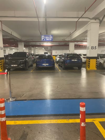 The image depicts an indoor parking garage, likely in Yunus Emre, Turkey, as indicated by the text on a car's license plate. The scene is well-lit by artificial lighting. The primary subjects are several parked cars, including a black SUV with the license plate "45 AR 393" in the foreground, a blue SUV with license plate "45 AOM 567" behind it, and a blue car with license plate "45 AVD 137" further back. The parking spaces are marked with yellow lines on the concrete floor, and there are blue and yellow stripes forming a lane.
Notable details include various signs and markings within the garage. On a white pillar, the text "B5 B5" is clearly visible. Another pillar has the text "B4" and "C4" and "D4". A blue sign with white text reads "ENGELLİ ARAÇ PARK YERİ", which translates to "Disabled Vehicle Parking Area". There are also red pipes running along the ceiling and walls, typical of fire suppression systems.
In the foreground, a red and white striped bollard is positioned on the right side of the image. The overall atmosphere is that of a functional, everyday parking facility. There are no people visible in the image, and the activity is simply cars parked in their designated spots. The time of day cannot be determined, but the artificial lighting suggests it could be day or night. The weather is not relevant as it is an indoor location.