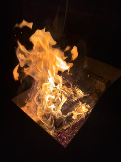 A close-up, angled shot shows a metal fire pit containing flames consuming wood. The pit's edges are visible as dark, slightly corroded metal. Inside, flames rise, with a mix of orange and white hues. Unburnt wood is visible in the background, along with dark charcoal and slight red embers. The background surrounding the fire pit is a solid, stark black. The location, Mayak, Russia, is not evident from the visual context.