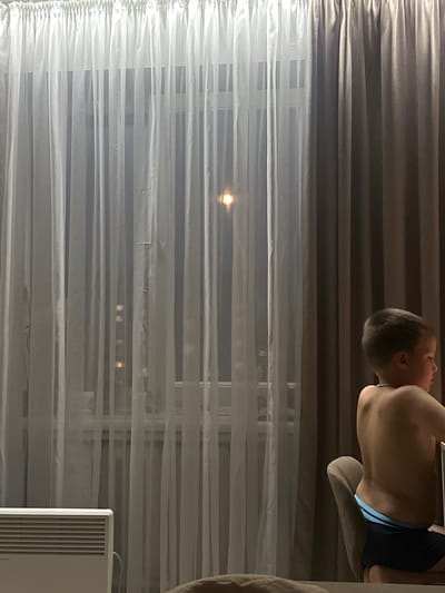 The image shows a young boy, viewed from the side and from behind, lying on his stomach. He is wearing blue swim trunks. He is positioned on a light-colored surface, possibly an armchair or a sofa, against a backdrop of dark, horizontal striped curtains. In front of the curtains, there are sheer white curtains covering a window. Through the sheer curtains, faint reflections of lights can be seen, suggesting it is evening or nighttime. The window panes are visible through the sheer fabric, and some horizontal blinds appear to be behind the sheer curtains. To the right of the image, a portion of a white electric heater with vertical slats is visible. The boy's body is arched backward, and his head is turned to the right, looking towards something out of frame. His expression is not discernible. The overall lighting is dim, contributing to a sense of interior evening. The setting appears to be a home environment.