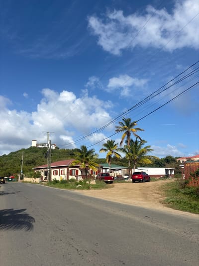 The image displays a daytime, roadside scene in Spanish Town, British Virgin Islands, under a blue sky with scattered white cumulus clouds. A paved asphalt road, with palm frond shadows on its left side, extends from the lower left towards the mid-right. A dirt shoulder runs alongside the road on the right. In the midground, a single-story building with a red roof, light-colored walls, and red shutters is situated on the right. A dirt lot in front of this building contains a red pickup truck and a red SUV. Several palm trees are distributed among the structures. Utility poles and multiple overhead power lines are visible across the upper midground. On the far left, a dark-colored vehicle is partially visible on the road. A prominent, heavily vegetated hill rises in the background, topped by a multi-story, light-colored building. No individuals are present or engaged in visible actions.