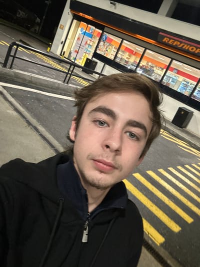 A young man with blue eyes and light brown hair poses for a selfie outdoors at night. He wears a dark hooded jacket, partially zipped. Behind him is a gas station convenience store with bright interior lights, visible through large windows. Yellow striped road markings are on the ground to his right, and a trash can is visible near the store. The overall lighting is dim and artificial.