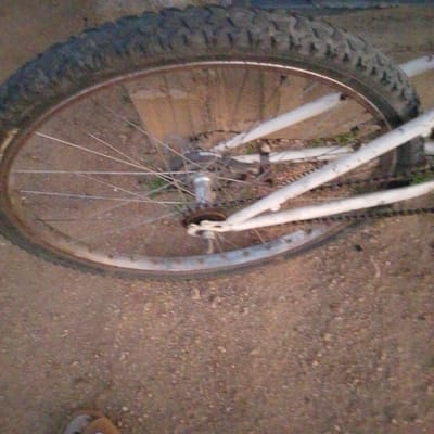 The image shows a close-up of a bicycle wheel with a knobby tire, spokes, and rim. The bicycle frame, painted white, is partially visible. The wheel is on the ground, which appears to be a dirt or gravel surface. The background is out of focus but suggests an outdoor environment. A person's shoe is visible at the bottom left corner of the image. The scene is set in Wuro Allahamdu, Nigeria.
