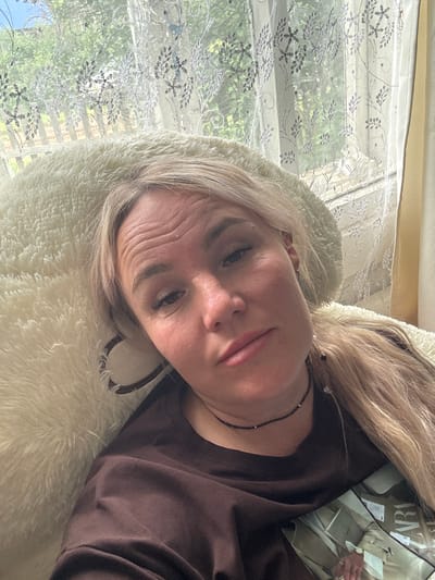A blonde woman with her hair in a ponytail is lying down, resting her head on a cream-colored fluffy pillow. She is wearing a dark brown t-shirt and a black beaded necklace. She is looking at the camera with a slight smile. Behind her, sheer curtains with a floral pattern are visible, allowing sunlight to filter through and illuminate the room. Outside the window, green foliage and a fence can be seen, suggesting an outdoor environment. A magazine is partially visible in the lower right corner of the frame, with the word "LARA" clearly visible. The image appears to have been taken indoors, likely in a residential setting. There is no information available about the location being in Pozdyshki, Russia.