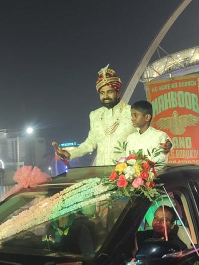 A groom in a decorated turban and traditional attire stands on a car, accompanied by a young boy dressed similarly. The car is adorned with flowers and garlands. A man is visible inside the car, possibly the driver. In the background, a banner with "MAHBOOB" is displayed, along with what appears to be an architectural structure and building. The event seems to be a wedding procession.