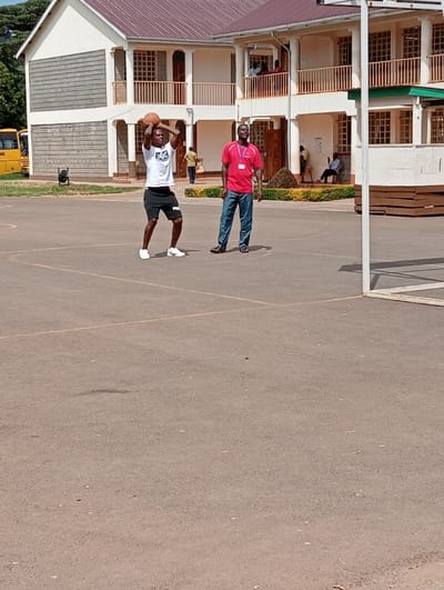 This image captures an outdoor basketball court scene, likely at a school or community center in Riara Ridge, Kenya. In the foreground, two adult males are engaged in a basketball activity under bright daylight conditions.
On the left, a dark-skinned man is captured mid-action, preparing to shoot a basketball. He is wearing a white short-sleeved t-shirt with a graphic design, black shorts featuring the "FILA" logo in white on the left leg, and white sneakers. He holds the basketball above his head with both hands, his body poised for a shot.
To his right, a second dark-skinned man stands observing. He wears a red short-sleeved polo shirt, dark blue jeans, and dark sandals. He is looking upwards and slightly to his left, seemingly watching the first man's actions or the unseen basketball hoop. A small, unidentifiable logo or ID badge is visible on his red shirt.
The background features a multi-story building with light-colored walls, interspersed with sections of grey-textured material, and topped with a reddish-brown roof. The building has numerous windows, some appearing to have security bars, and a second-story balcony where several people are visible. Green landscaping, including bushes, is present along the base of the building. On the far left, part of a yellow school bus with the word "SCHOOL" partially visible is parked. A white basketball hoop post is visible on the far right, with the hoop itself out of frame. The court itself is asphalt or concrete, with faded yellow lines marking its boundaries. The overall impression is one of casual recreation or practice on a sunny day.