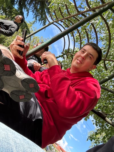 A group of people is gathered outdoors in Topoli, Bulgaria, under a green metal trellis with foliage overhead. The sky is blue with scattered clouds. One person, wearing a red hooded sweatshirt, is prominently featured in the foreground. They are holding a black smartphone. The sole of a white and grey shoe with red accents is visible close to the camera, obscuring part of the view. Two other individuals are partially visible in the background. One is wearing glasses and a black hoodie. The other is partially behind the person in the red sweatshirt. A building with a red roof is visible in the distance, as well as green trees.