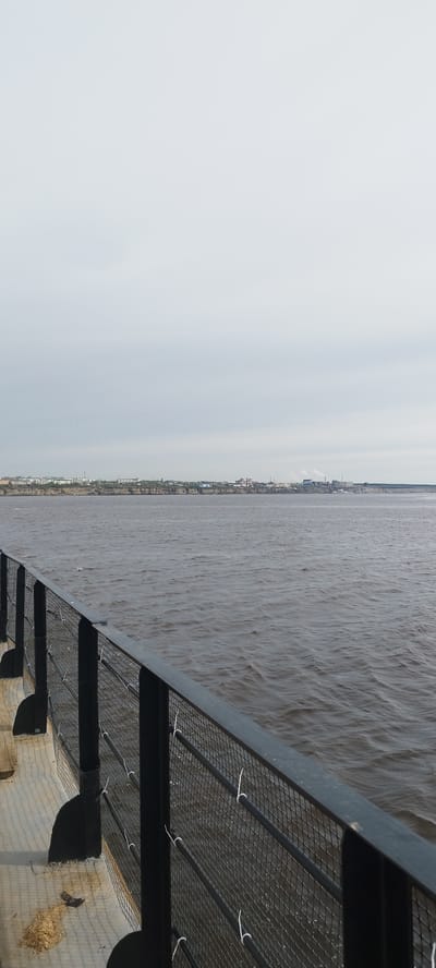 The image shows a view of a body of water, possibly a river, from behind a black metal fence. The water is dark and slightly choppy. On the opposite bank, a cityscape is visible with buildings and industrial structures, including what appear to be factories with smokestacks. The sky is overcast and gray. The fence is in the foreground and appears to be part of a walkway or deck. The white ties that are connected to the fence are holding something in place, but it is unidentifiable in the image. Given the description of Bestyakh, Russia, this is likely a view of the Lena River.