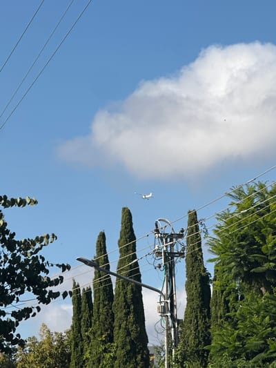 The image shows a blue sky with a large white cloud on the left side. Power lines crisscross the lower left and upper right portions of the frame. An electrical pole with various components is visible in the upper right. Trees are on the right side of the image. A small white object, potentially a kite or a drone, is in the sky between the cloud and the electrical pole. There are also several small black objects in the sky, potentially insects. The location is identified as Kfar Shmaryahu, Israel.