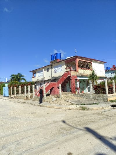 A daytime exterior photograph depicts a two-story residential building located in Ciego de Ávila, Cuba. The environment features a clear blue sky with minimal clouds and an unpaved, gravelly dirt road in the foreground.
The building is painted in a pale yellow or cream hue and includes a prominent external red concrete staircase leading directly to a second-story balcony. The balcony features a red railing and yellow-colored decorative metal grating. Two blue plastic water tanks are visible on the flat roof, alongside a television antenna and a satellite dish. Windows on both levels are fitted with white louvered shutters. A portion of the ground floor wall beneath the staircase displays a lighter, possibly blue or green, color.
A low fence constructed of concrete posts borders the property, behind which red flowering bushes are cultivated. A blue metal gate is partially visible to the left of the main structure.
One individual, dressed in dark long-sleeved attire, stands on the unpaved shoulder next to the road, near the property's fence line. The person is positioned facing generally towards the building's facade. Strong sunlight creates distinct shadows across the road, including a long, narrow shadow originating from the right side of the frame, and a shadow cast by the individual.