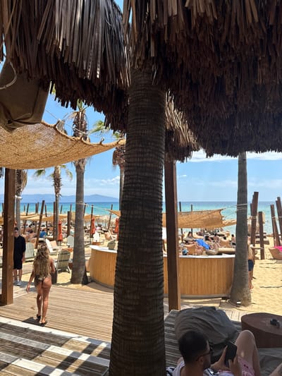 The image depicts a beachside establishment in Nea Peramos, Greece, characterized by a thatched roof structure providing shade over a wooden deck and sandy beach area. The scene is populated with numerous beachgoers relaxing on lounge chairs and under umbrellas. Palm trees are prominently featured, with their fronds contributing to the tropical ambiance. The ocean is visible in the background, meeting a clear blue sky with distant hills. In the foreground, a woman with long blonde hair is walking away from the camera on the wooden deck, wearing a dark bikini. Another person, a man in a black shirt and shorts, is walking towards the left edge of the frame. Further back, several people are seated on lounge chairs and beanbag chairs. A wooden bar area is visible behind the seated patrons. The overall atmosphere suggests a leisurely day at a beach resort.