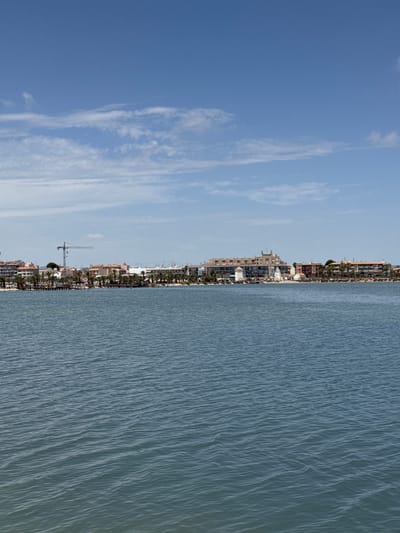 The image shows a body of water in the foreground, with a town situated along the opposite shore. The town consists of low-rise buildings, some with balconies and tiled roofs, interspersed with palm trees. A prominent feature in the town is a construction crane, suggesting ongoing development or renovation. In the distance, the sky is blue with scattered white clouds. The water appears calm with small ripples. The location is identified as the waterfront of San Pedro del Pinatar, Spain, likely overlooking the Mar Menor lagoon.
