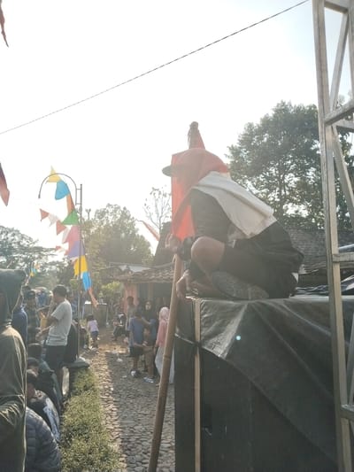 The image shows an outdoor gathering with a crowd of people. In the foreground, a person is crouched down on top of a black tarp-covered object, wearing dark clothing and a white cloth draped over their shoulders. Behind this individual, colorful triangular flags are strung on poles, and a larger red flag is visible. A crowd of onlookers is gathered on a cobblestone path lined with greenery. In the background, there are buildings with tiled roofs and trees. The scene appears to be set during daylight, with bright sunlight casting shadows. The atmosphere suggests a community event or celebration. The context of Ngadirejo, Indonesia, implies this could be a local festival, market, or cultural event taking place in a village or small town setting.
