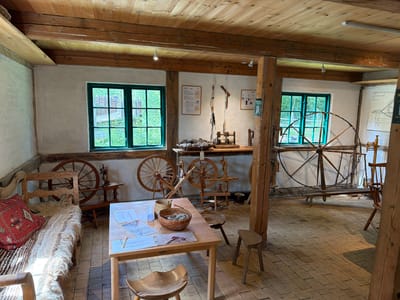 This is an interior photograph of a room in the city of Lejre, Denmark, likely a museum exhibit. The room features a wooden-beamed ceiling and white-washed walls, with a brick-patterned tiled floor. Several large, traditional spinning wheels are visible, some standing upright and others laid horizontally. In the foreground, a wooden table displays papers and baskets containing what appears to be wool or fiber. To the left, a rustic wooden bench is covered with a textured blanket and a red cushion. Natural light enters through two large, green-framed windows, revealing greenery outside. The overall impression is one of a historical craft space, possibly a workshop or exhibition dedicated to textile production. No people are visible in the image.
