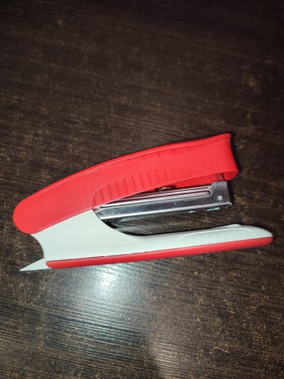 A red and white stapler rests on a dark wood surface. The stapler is positioned at an angle, with its top red section slightly raised, revealing the metallic stapling mechanism. The bottom white section, trimmed with red, forms the base. The wood surface exhibits a distinct grain pattern, with visible lines and some lighter-colored speckles. The lighting appears to be overhead, casting subtle shadows and highlighting the texture of both the stapler and the wood. There are no people or other objects in the frame. The image does not provide any information that would allow for its location in the city of Ivanovka, Russia, or any other specific geographical location.