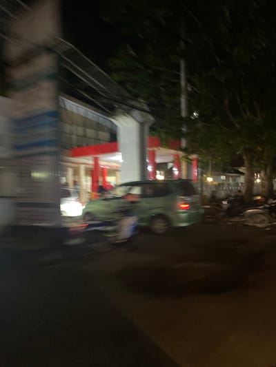 A nighttime, blurry image shows a street scene in Walennae, Indonesia. A light green van is parked on the right side of the street, its brake lights illuminated. Behind the van, several motorcycles are parked. A building with red trim and white pillars is visible on the left side of the street. Trees line the right side of the street, and lights are visible in the windows of buildings in the background. The image is out of focus, suggesting it was taken with a handheld camera while moving.