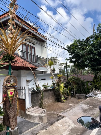 The image depicts a residential area in Susut, Indonesia, with a focus on a dwelling featuring a white facade and a red-tiled roof. A prominent element in the foreground is a decorative pole draped with brown fabric, adorned with a basket containing offerings. Adjacent to the house, a balcony with a black railing is visible, and above, a network of black utility wires crisscrosses the sky. The scene is filled with lush greenery, including trees and various potted plants. The ground is a mix of dirt, gravel, and concrete pathways. A motorcycle mirror is visible in the lower right corner, reflecting trees and a person. The overall impression is one of a typical tropical environment with elements of local culture evident in the decorative offerings.