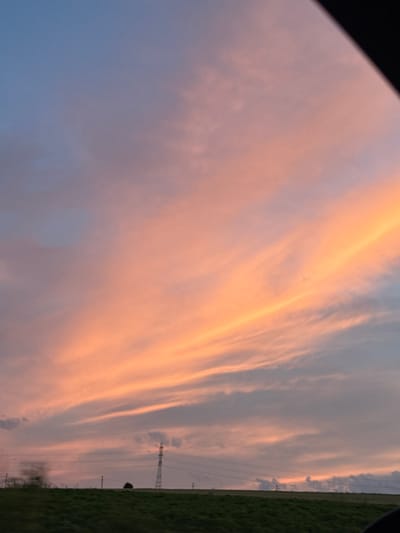 The image depicts a rural landscape under a vibrant sunset sky. The sky is filled with wispy clouds painted in hues of orange, pink, and blue. In the distance, a tall, metallic electricity pylon stands prominently against the horizon, suggesting the presence of power infrastructure in the area. A dark, solitary tree is visible to the left of the pylon. The ground is covered in green grass, indicating a natural or agricultural setting. The scene appears to be captured from a moving vehicle, as evidenced by the slightly blurred foreground and the dark frame in the upper right corner of the image, which could be part of the vehicle's interior. The image does not contain any people or specific urban features that would pinpoint its location within Zvegor, Bulgaria, but it conveys a sense of open, undeveloped land bathed in the warm light of dusk.
