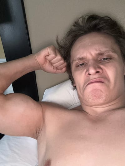 The image shows a close-up, selfie-style shot of a shirtless man in bed. He is lying on his back, with his left arm flexed, showcasing his bicep. His face is contorted in an exaggerated expression, with his eyebrows furrowed and his mouth pursed. The background appears to be a bed with white sheets and a neutral-colored wall or headboard. There are no discernible landmarks or indicators of a specific location in San Francisco, United States of America. The lighting suggests an indoor setting, possibly during the day given the natural skin tone.