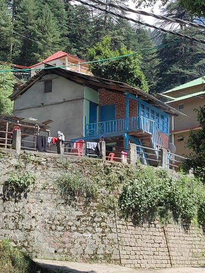 In the city of Chhyal, India, a two-story residential building is depicted, nestled amongst a dense forest of evergreen trees. The building features a rough stucco exterior on its left side and exposed brickwork on the right, with a weathered tin roof. A vibrant blue wooden balcony with an attached staircase overlooks the front of the house, accessible from the upper level. Clothes are hung to dry on a railing that extends across the lower front portion of the property. Two red plastic chairs and a small satellite dish are also visible. The house is situated on a steep slope, supported by a retaining wall constructed from rough-hewn stones. Overgrown green vegetation covers the wall and the surrounding area. In the background, a portion of another building with a red roof and white trim can be seen peeking out from behind the trees. The sky is partly obscured by a network of overhead wires.