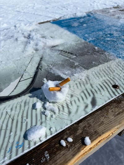 The scene depicts a small, crude snowman crafted from snow, approximately 5-7 cm in diameter, situated on an outdoor information panel within the ski resort area of La Clusaz, France. Two orange-filtered cigarette butts are embedded vertically into the snow sphere. The panel surface immediately surrounding the snow figure is a textured, pale greenish-white material exhibiting multiple fine, dark linear scratches and a single dark circular fixture to the right. This panel transitions into a surface covered by a thin layer of ice and snow, beneath which a graphic or map is discernible, featuring dark linear patterns and blue sections, indicative of a ski resort map. The background consists entirely of undisturbed white snow, typical of a ski slope or open snowy area, under bright daylight conditions. To the right, a weathered wooden plank, likely part of a bench or railing, is also partially snow-dusted. No individuals are present.