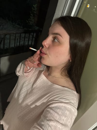 A young woman is holding a lit cigarette to her lips. She has long brown hair, light skin, and wears a light pink patterned shirt. She is wearing a necklace and ring. The background shows a window and what appears to be a dark outdoor space with some greenery. The city of Chornomorskoe, Ukraine, is not evident in the image.