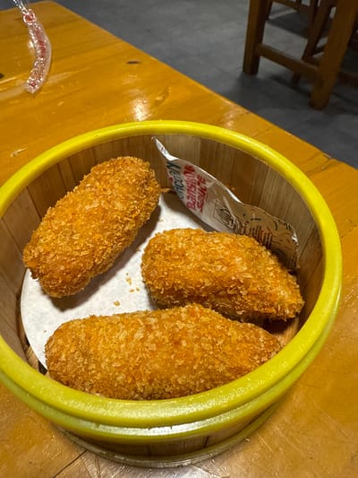 A close-up shot captures a bamboo steamer basket filled with three golden-brown, breaded items. These items appear to be fried snacks, possibly croquettes or spring rolls, arranged on a white paper liner within the steamer. A small packet of "Bangkok Sauce" is partially visible, peeking out from under one of the fried items. The steamer basket itself has a bright yellow rim. The entire basket rests on a warm-toned wooden table. In the background, a portion of a wooden chair or table leg is visible, suggesting a dining setting. The lighting is warm and indoor, casting soft shadows and highlighting the crispy texture of the food. The overall impression is one of a casual dining experience, likely a meal or appetizer. The visible text on the sauce packet includes "Bangkok Sauce" and what appears to be a halal certification logo.