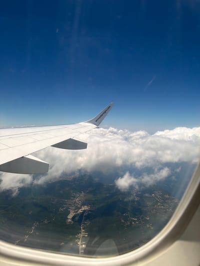 An aerial view from an airplane window shows a white airplane wing extending into a bright blue sky. Below the wing, white clouds partially obscure a lush green mountainous landscape dotted with buildings and roads, suggesting a town or city. The text "airmontenegro.com" is visible on the wing. The scene appears to be over Kubasi, Montenegro.