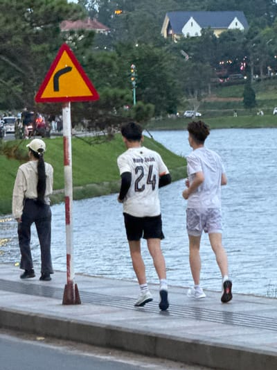 The image captures a view along Xuan Huong Lake in Da Lat, Vietnam. In the foreground, three individuals are seen from behind. The person on the left is wearing a light-colored jacket, dark pants, and a white cap with a long dark ponytail. They are standing with their back to the camera. To their right, a person in a white t-shirt with the number "24" and black sleeves, and black shorts is jogging. Further to the right, another person in a light-colored t-shirt and patterned shorts is also jogging. A traffic sign indicating a sharp curve to the left is prominently displayed on a pole in the middle ground. In the background, Xuan Huong Lake is visible, with a grassy embankment and trees lining the opposite shore. Buildings, including a house with a blue roof, are nestled amongst the trees on a hill. Several vehicles are present on the road in the distance. The overall scene depicts a leisurely outdoor activity by the lake.