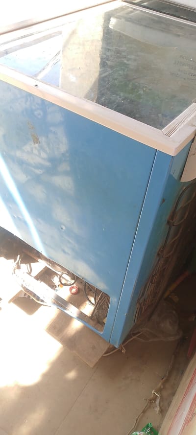 The image displays a used, light blue chest freezer with a clear glass top, situated in an outdoor or semi-outdoor setting in Danbare, Nigeria. No people are visible.

The freezer's exterior shows signs of wear, including several dents on its blue metallic sides. The glass lid appears somewhat scratched and dirty, reflecting bright sunlight. Faint, inverted text is visible on the underside of the glass, possibly a brand name or product information, with words like "WORLD" and "FREEZ..." partially discernible.

The scene is brightly lit by strong sunshine, suggesting it is daytime. On the left side of the freezer, the lower panel is open, exposing internal machinery, wires, and what appears to be a compressor unit. A small red cap or object is located among these components. A wooden or cardboard shim is placed under the front-left corner of the freezer, slightly elevating it from the concrete floor.

To the right of the freezer, cables are visible, likely connected to the unit. Further right, a dark, grid-like structure, possibly a vent or part of the cooling system, is present. The floor is concrete, and in the bottom right corner, a green plastic bag and other debris are scattered. The overall impression is a functional, utilitarian space, possibly a small market stall or storage area, with equipment showing extensive use.