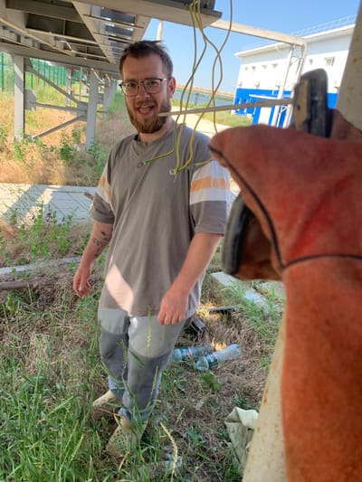 A man with glasses and a beard stands outdoors, partially obscured by an orange glove in the foreground. He is wearing a grey t-shirt with orange and white stripes on the sleeves and grey pants. He is smiling and holding yellow and green wires. The background includes tall grass, metal structures, a building with blue and white accents, and a fence. The scene appears to be in Pivdennoukrainsk, Ukraine, likely near industrial infrastructure given the metal structures and building. Two plastic bottles are visible on the ground. The lighting suggests it is daytime.