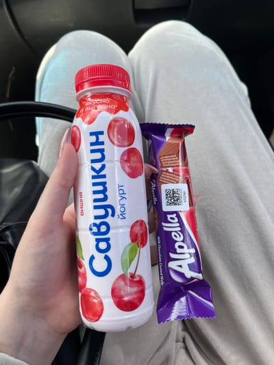 A close-up shot shows a person's hand holding a chocolate bar and a bottle of cherry-flavored yogurt. The chocolate bar, a "Alpella Milk Chocolate Coated Wafer," is purple and red with an image of the wafer inside. The yogurt bottle is white with a red cap and features an illustration of cherries. The text on the yogurt bottle is in Russian and reads "вкусно и выгодно" which translates to "tasty and profitable," and "черный" which translates to "black" or "cherry." The scene appears to be indoors, possibly in a car, given the dark background and the texture of the clothing, which looks like white sweatpants. The lighting suggests it is daytime. The person's fingernails are painted a shimmery pink. The overall impression is a casual moment of someone holding snacks.