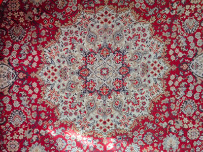 The image shows a close-up view of a red and white oriental rug with a complex floral pattern. The rug has a central medallion design and is surrounded by a border of smaller floral motifs. The colors are predominantly red, white, and gray, with some accents of blue and brown. The lighting suggests the rug is indoors, possibly in a home. There is no indication of the city of Nadezhda, Russia, or any specific location within it.