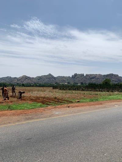 The image shows a rural scene in Makera, Nigeria. A paved road with a yellow line runs along the right side of the frame. Adjacent to the road is a strip of red soil and green vegetation. Further to the left, the landscape transitions into a field, with a row of thin trees. In the foreground, several people are observed standing in the field. In the background, rocky hills are visible under a partly cloudy sky.