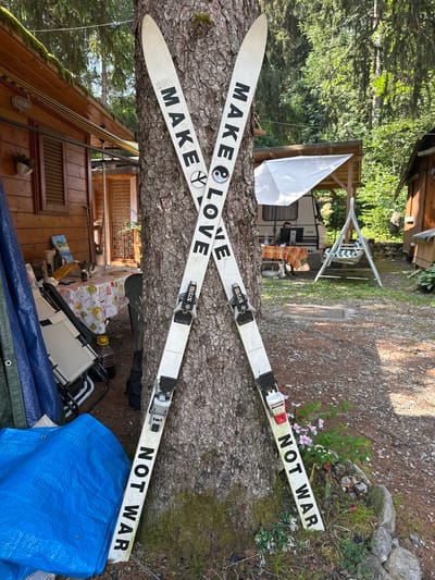 Two skis are propped against a tree, crossed in an X formation. The skis are white with black text. The left ski reads "MAKE LOVE" with a peace symbol and a yin-yang symbol. The right ski reads "NOT WAR". The skis are positioned in an outdoor setting, likely a campsite. Behind the skis, a wooden cabin with a red-striped awning is visible, along with a camper trailer and a white swing set. The ground is covered in gravel and dirt, with some sparse vegetation around the base of the tree and in front of the right ski. The image appears to be taken during the daytime, with sunlight filtering through the trees. The location is identified as Borno, Italy. There are no people visible in the frame.