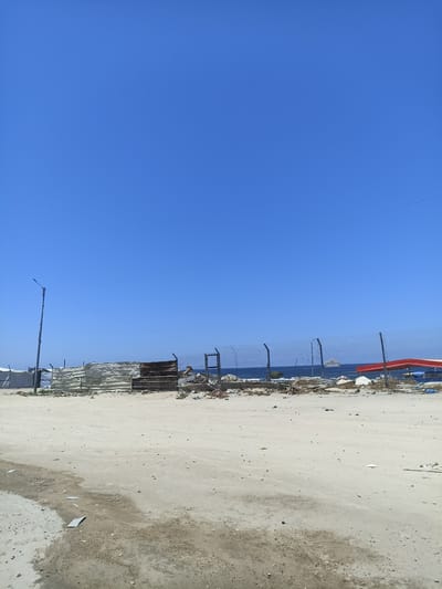 This media file depicts an outdoor scene under a clear blue sky. The foreground is a sandy, uneven ground with some scattered debris and puddles. A makeshift fence or barrier made of corrugated metal and wood runs horizontally across the mid-ground, separating the sandy area from what appears to be a body of water in the background. Beyond the fence, a large expanse of blue water is visible, with a small island or rock formation in the distance. A wire fence with metal posts also runs along the water's edge, and there are some structures or tents behind the corrugated metal barrier, some appearing to be made of white canvas. A tall light pole is situated to the left of the frame. The overall impression is one of a somewhat desolate or underdeveloped coastal area, possibly undergoing construction or in a state of disrepair. The clear sky and bright sunlight suggest it is daytime, likely a warm and sunny day. There is no visible text in the image.