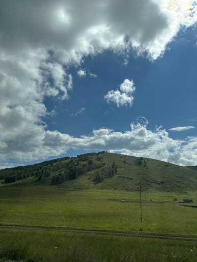 The image depicts a vast green meadow in front of a rolling, tree-covered hill. The sky above is a vibrant blue, dotted with large, fluffy white clouds, with the sun peeking through some of them. In the mid-ground of the meadow, there are several wooden structures, possibly pens or sheds, and a single utility pole. A dirt path or road is visible in the foreground, running horizontally across the frame. The overall impression is one of a rural, natural landscape bathed in sunlight. The location is not specified as being in the city of Uralsk, Russia.