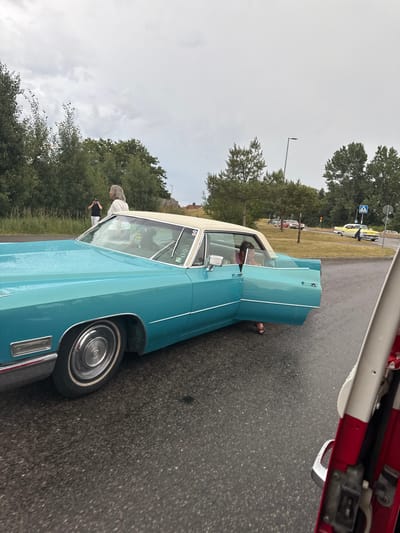 The image depicts a cloudy day in Falkenberg, Sweden, likely during an event or gathering related to vintage cars. The foreground features a turquoise Cadillac with its driver's side door open. A person with dark hair, wearing a pink shirt, is seated in the driver's seat, looking towards the front. To the left of the Cadillac, a woman with blonde hair, wearing a white top, is standing and holding what appears to be a camera. Further in the background, another person in a dark shirt and white pants is visible. Several other classic cars, including a yellow and a red one, are parked or driving in the distance on a wet asphalt road. Trees and vegetation line the background, suggesting a location near a park or a residential area. The overall scene captures a moment during an outdoor car exhibition or cruise.
