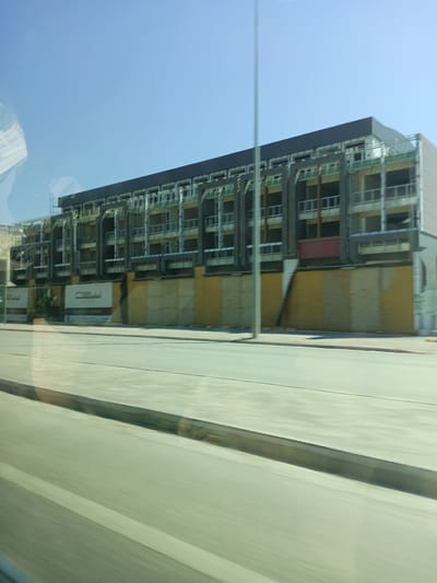 This media file depicts a large, multi-story building under construction, viewed from a slight elevation. The building is dark gray and has a linear, modern design with repeating balcony structures. Many of the window openings are visible as dark voids. The building is covered in scaffolding and construction materials, indicating an ongoing project.
The lower facade of the building is partially covered by large, light-colored panels, some with yellow accents. To the left, there are two visible signs. One is white with dark text that appears to say "new". The other sign, also white, has text that is difficult to decipher due to resolution. A small amount of greenery is visible between the building and a wooden fence.
In the foreground, a paved road or walkway runs horizontally across the image, with a distinct curb. The surface of the road is blurred due to motion, suggesting the image was taken from a moving vehicle. The sky is a clear, bright blue, indicating a sunny day with no clouds. A tall, thin pole stands vertically in front of the building, partially obscuring it. The overall scene suggests an urban or suburban setting where a significant construction project is underway. The time of day appears to be daytime, likely late morning or early afternoon, given the bright sunlight and clear sky.