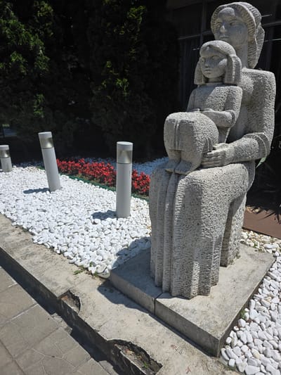 This media file depicts a stone sculpture of three figures, possibly a family, huddled together. The figures are rendered in a rough, textured style, with their faces and bodies showing a sense of communal embrace and perhaps melancholy. The sculpture is situated outdoors, with a background of dark green foliage, possibly a hedge or trees. In front of the sculpture, there is an area covered in white pebbles, with a line of red flowers bordering a grassy area. Three cylindrical, white lights with translucent tops are partially embedded in the ground amidst the white pebbles. The ground surrounding the sculpture is concrete, with some paving stones visible in the foreground. The lighting suggests it is daytime, with shadows indicating bright sunlight. The scene appears to be a public space, possibly a park or garden, in Dimovo, Bulgaria, given the provided location context. No visible text is present in the image. The overall mood conveyed by the sculpture is one of intimacy and possibly hardship or contemplation.