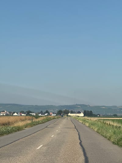 A road bends to the right, its asphalt cracked and marked with white lines. To the right, a grassy verge separates the road from a landscape with dense vegetation. Houses and buildings line the roadside, leading towards a distant village or town, indistinct due to atmospheric haze. The sky, a clear gradient of blue, fills the left half of the scene. A tall communication tower is visible in the middle ground.