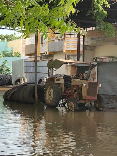 The image depicts a flooded scene in the city of Malkapur, India, likely an industrial or commercial area given the presence of a tractor, stacked tires, and buildings with signage. A light-colored tractor, with a red front grille and a canvas canopy, is partially submerged in murky floodwaters. Several large tires, some stacked and others leaning, are also in the water, some appearing to be used tractor or vehicle tires. In the background, a building with beige and orange striped sections is visible, along with corrugated metal structures. One building has a sign that reads "ओम गुड्स ट्रांसपोर्ट सर्विस" (Om Goods Transport Service), suggesting a business operation. The ground is uneven and appears to be a mixture of mud and water. Green foliage from a tree overhangs the scene from the top of the image, casting shadows. The overall impression is one of disarray and the impact of heavy rainfall or flooding.