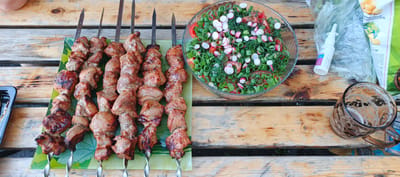 The image depicts an outdoor setting, likely a picnic or barbecue, on a wooden table. In the foreground, several skewers of grilled meat are arranged on a green, leaf-patterned platter. To the right of the meat, a glass bowl is filled with a fresh salad composed of chopped tomatoes, radishes, and green onions, garnished with dill. A clear glass mug, containing a foamy beverage, is positioned on the right side of the table. In the background, a white nasal spray bottle and a plastic bag containing what appear to be potato chips are visible, suggesting a casual meal. The wooden planks of the table show signs of wear, and the overall scene evokes a relaxed, outdoor dining experience. The presence of Russian text on the packaging further indicates the location. Without specific landmarks, it is impossible to pinpoint the exact location within Petukhovo, Russia, but the scene is consistent with a typical outdoor gathering in a Russian context.