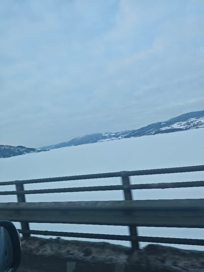 The image depicts a winter landscape captured from a moving vehicle on a bridge or elevated road in the vicinity of Moelv, Norway. In the foreground, a dark gray metal bridge barrier, composed of multiple horizontal rails and vertical supports, stretches across the lower half of the frame. A blurred, dark-colored vehicle side mirror is partially visible in the bottom-left corner. Beyond the barrier, a vast, flat expanse of white snow covers a frozen body of water, likely Mjøsa. The distant horizon is defined by snow-dusted mountains and hills, featuring patches of dark conifer trees and scattered structures on their slopes. The sky is uniformly overcast with light gray clouds, indicating a cold, winter day. The slight motion blur on the barrier is consistent with the photograph being taken from a moving vehicle. No individuals are visible.
