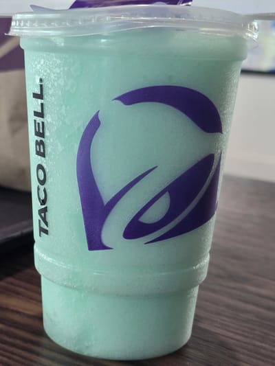 The image shows a close-up of a light green frozen beverage in a clear plastic cup with the Taco Bell logo. The cup is partially filled, with condensation visible on the exterior. The beverage appears to be frothy and icy. The Taco Bell logo, in purple, is prominently displayed on the side of the cup. The cup is sitting on a wooden surface, possibly a table, with a blurred background. There are no people or other distinct objects visible in the scene. The overall impression is of a refreshing drink, likely from Taco Bell. The location of Roswell, United States, is not evident from the image itself.