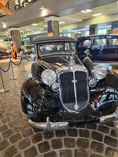 The image shows a black vintage car with a large, shiny chrome grill and headlights, situated inside a building with a brick floor. The building appears to be a museum or exhibit space. The car is near a roped-off area, likely to prevent visitors from touching it. In the background, there are other exhibits and historical vehicles. The lighting is bright, illuminating the car and other objects in the museum. The location is in the city of Архангельское, Russia.