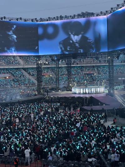 The image depicts a concert venue, likely a stadium, filled with a large crowd of people. The venue appears to be located in Goyang-si, South Korea, as indicated by a sign in the image. The crowd is holding up illuminated objects, possibly light sticks or phones with lit screens, creating a sea of light. A large screen displays an image of a person. A circular stage is present on which people are standing.